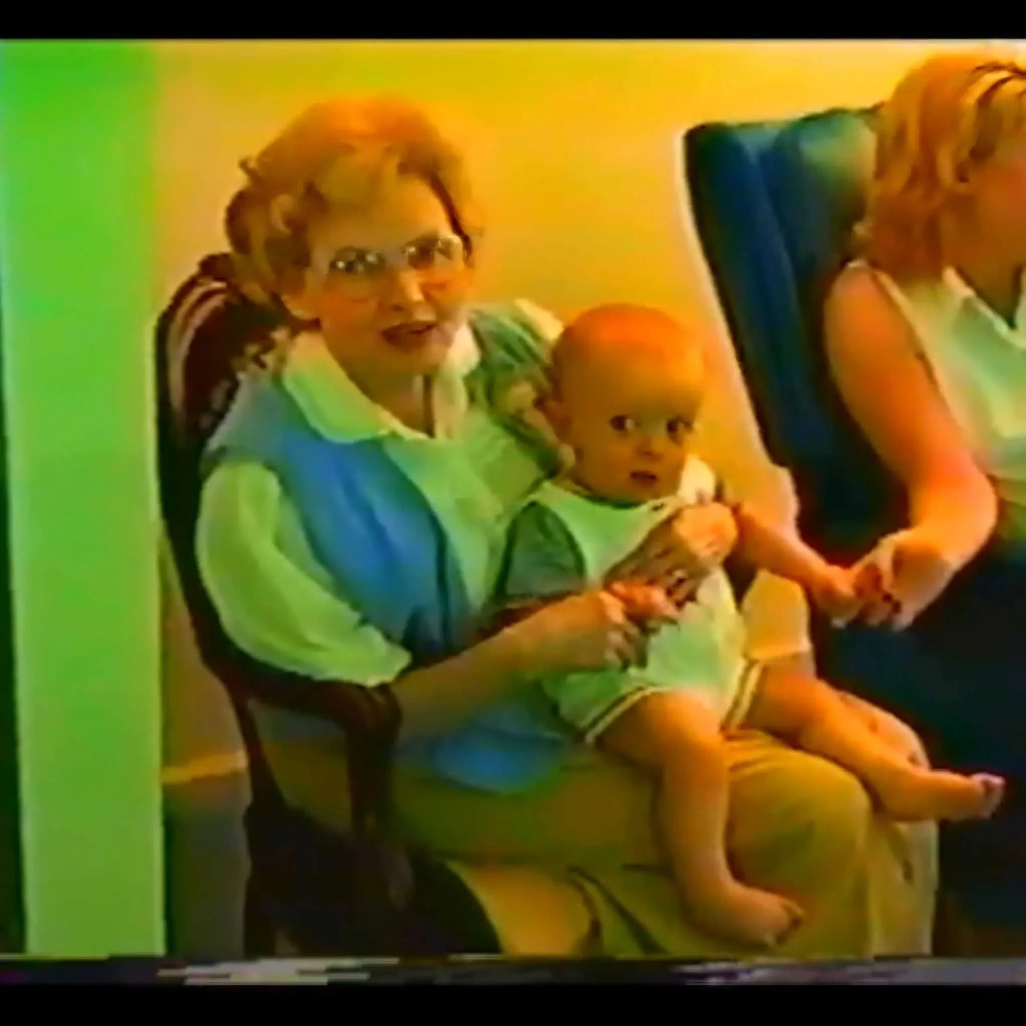 VHS frame of Gruzel seated indoors holding baby Caleb